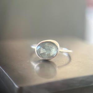 May include: A silver ring featuring a faceted, oval-shaped, pale green gemstone set in a smooth metal bezel. The stone displays internal inclusions and a translucent quality, while the thin, polished silver band completes the design.