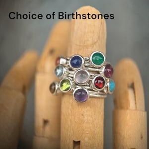 Könnte beinhalten: Ein stapelbarer Silberring mit mehreren runden, im Cabochon-Schliff geschliffenen Edelsteinen in verschiedenen Farben wie Blau, Grün, Rot, Violett und Transparent. Der Text "Choice of Birthstones" ist über dem Ring zu sehen.