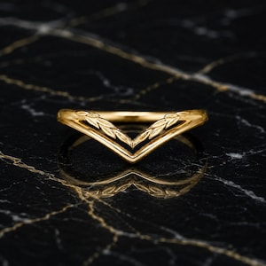 Peut inclure: Une bague en or en forme de V avec un motif de feuilles gravé délicat sur la bande supérieure. La bague présente une finition métallique polie et un design en chevron pointu qui crée une silhouette symétrique, reflétant la lumière.