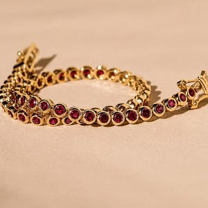 Könnte beinhalten: Ein goldenes Tennisarmband mit einer durchgehenden Reihe runder, in Zargenfassung gefasster roter Rubine. Das Armband besteht aus polierten Goldgliedern und verfügt über einen sicheren Kastenschloss-Verschluss, der ein klassisches Design bietet.