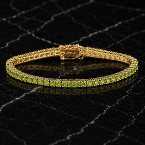 May include: A gold-toned tennis bracelet featuring a continuous row of round, vibrant lime-green peridot gemstones set in a metal frame. The bracelet includes a secure box clasp mechanism with a safety latch, creating a uniform circular design.