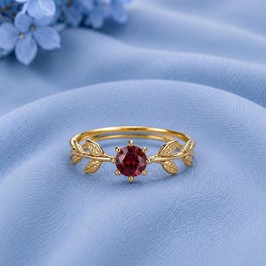 May include: A gold ring featuring a round, deep red gemstone at the centre, held by a six-prong setting. The band is designed with delicate, symmetrical leaf motifs extending from the central stone along both sides of the circular ring.