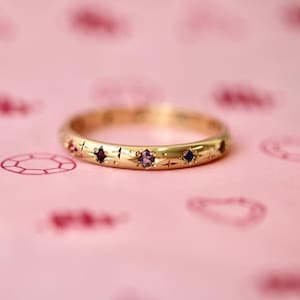 Op de afbeelding: Een gouden ring met een reeks kleine, ronde, veelkleurige edelstenen die in het metaal zijn gezet. Het oppervlak van de gouden ring is versierd met gegraveerde sterpatronen rondom elk van de ingelegde, kleurrijke, ronde stenen.