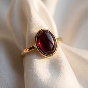 May include: A gold ring featuring a smooth, oval-shaped deep red cabochon gemstone set in a polished metal bezel. The band is thin and circular, crafted from a reflective gold-toned metal that catches the light, highlighting the dark red stone.