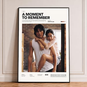 May include: A framed movie poster for A Moment to Remember, featuring a central photograph of a person giving another person a piggyback ride. The poster includes the film title, director John H. Lee, cast names, release date, and a PG-13 rating label.