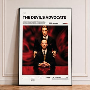 May include: A framed movie poster for The Devil's Advocate featuring a central image of two men in suits against a red background. The poster includes the film title, director Taylor Hackford, cast names, release date, and a restricted rating label.