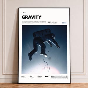 Może przedstawiać: Oprawiony plakat filmowy Gravity przedstawiający ciemnoniebieską scenę kosmiczną z astronautą unoszącym się w centrum. Biała ramka zawiera tytuł Gravity, reżysera Alfonso Cuaróna, obsadę, datę oraz oznaczenie wiekowe PG-13.