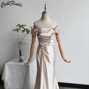 May include: A floor-length champagne-colored satin gown featuring an off-the-shoulder neckline with pleated fabric detailing. The dress includes floral embellishments on the left shoulder and waist, with a draped skirt and a subtle train.