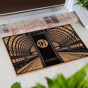 May include: A rectangular tan doormat with a black graphic print depicting a train station platform. The design features a central pillar with the text 9 3/4 inside a circle, flanked by arched ceilings and train carriages on tracks.