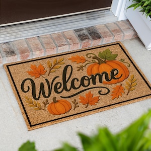 May include: A rectangular tan coir doormat featuring a black border and the word Welcome in a black cursive script. The design includes two orange pumpkins and various autumn leaves in shades of orange, yellow, and green scattered across the mat.