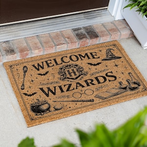 May include: A rectangular tan coir doormat featuring black printed graphics and the text "WELCOME WIZARDS". The design includes a central crest with an "H", a cauldron, a wand, round glasses, an open book, potion bottles, and a pointed hat.