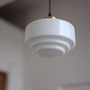 May include: A white glass pendant light fixture with a tiered, stepped circular design. The lamp features a smooth, glossy finish and is suspended by a thin black cord attached to a metallic gold-toned cap at the top of the glass shade.
