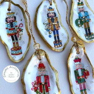 May include: Five oyster shell ornaments featuring hand-painted nutcracker figures in various uniforms, accented with small clear rhinestones. Each shell has a gold-painted edge and a gold hanging cord with a bead. Text reads Coastal Accessories by Jennifer Playford.