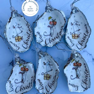 May include: Six oyster shell ornaments featuring hand-painted illustrations of mice with Christmas gifts and the text merry Christmas. Each shell has silver glitter edges, a silver hanging cord, and small clear rhinestones. Coastal Accessories by Jennifer Playford logo is visible.