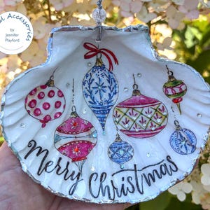May include: A white scallop shell ornament featuring hand-painted colourful Christmas baubles in shades of pink, blue, and green, accented with small clear rhinestones. The text Merry Christmas is written in black script at the bottom.