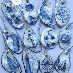 May include: A collection of twelve white oyster shell ornaments featuring blue nautical illustrations including lighthouses, whales, anchors, compasses, and sailboats. Each shell is accented with small clear rhinestones and hangs from a silver cord.
