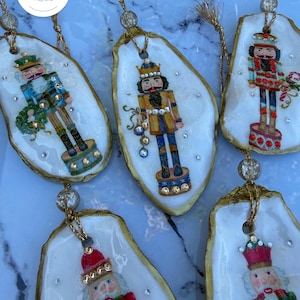 May include: Five oyster shell ornaments featuring hand-painted nutcracker figures in various uniforms. Each shell has a gold-painted edge, a gold hanging cord with a clear bead, and small embedded crystals. The nutcrackers hold items like wreaths.