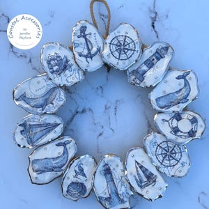 Peut inclure: Une couronne circulaire composée de coquilles d'huîtres individuelles, chacune ornée d'illustrations détaillées à l'encre bleue sur le thème marin, incluant phares, baleines, voiliers, ancres et boussoles, agrémentées de petites perles argentées.