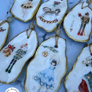 May include: A collection of oyster shell ornaments featuring hand-painted festive designs including a ballerina, rocking horse, nutcracker, and mouse king. Each shell has gold-painted edges, a gold hanging cord, and small decorative rhinestones.