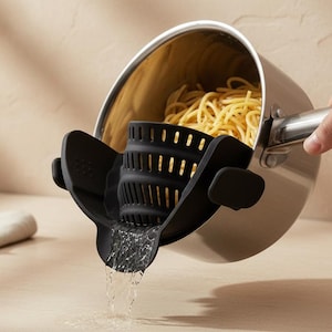 May include: A black silicone clip-on pan strainer attached to the rim of a stainless steel saucepan. The strainer features a curved, slotted design with a pouring spout, allowing water to drain while keeping cooked spaghetti inside the pan.