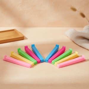 Könnte beinhalten: Ein Set aus zehn Kunststoff-Verschlussclips für Beutel, die in einem Halbkreis angeordnet sind. Die Clips sind lang, dünn und rechteckig mit einem Klappmechanismus und in leuchtenden Farben wie Pink, Gelb, Grün, Blau und Magenta gehalten.