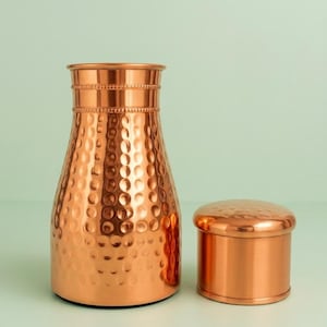 May include: A polished copper water pitcher with a hammered texture on the body and a smooth neck featuring a beaded rim, accompanied by a matching cylindrical copper lid with a slightly domed top and a smooth finish on the sides.