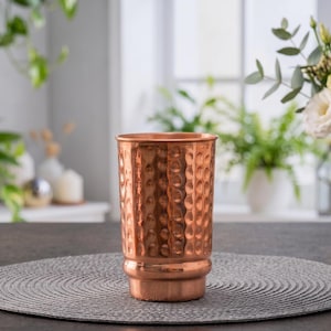 Puede incluir: Un vaso cilíndrico de cobre que presenta una textura martillada distintiva en toda su superficie exterior. El recipiente tiene un borde liso y ensanchado y una base ligeramente cónica, con un acabado metálico de color marrón rojizo.