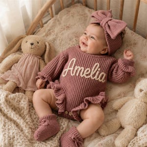 May include: A dusty rose knitted baby romper featuring the name Amelia embroidered in white script across the chest. The garment includes long sleeves with ruffled cuffs, a ruffled waistline, matching knitted booties, and a large bow headband.