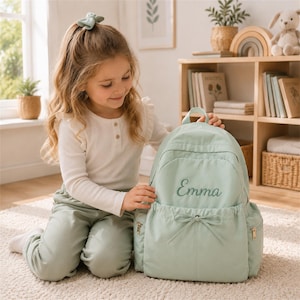 May include: A sage green backpack featuring the name Emma embroidered in a cursive script on the front pocket. The bag includes a drawstring closure with a bow detail, side pockets with gold-toned zippers, and a top handle for easy carrying.
