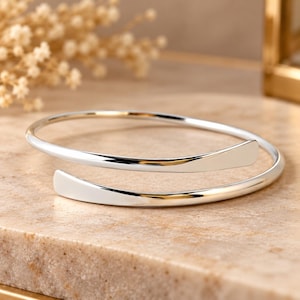 May include: A polished silver-tone bypass bangle bracelet featuring a smooth, reflective surface. The design consists of a single continuous band that wraps around to create an open, overlapping silhouette with two flattened, rounded ends.