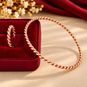 Op de afbeelding: Een roségouden armband met een gedraaid touwontwerp en een bijpassende ring met hetzelfde gedraaide touwpatroon. De ring staat rechtop in een rood fluwelen juwelendoosje, terwijl de ronde armband ernaast op een glad oppervlak ligt.