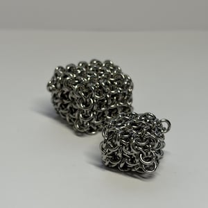 May include: Two three-dimensional cubes constructed from interlocking silver-coloured metal rings. The larger cube is positioned behind the smaller cube, both featuring a dense, woven chainmail pattern with a reflective, metallic finish.