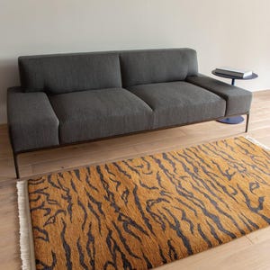 Könnte beinhalten: Ein rechteckiger Teppich mit Tigerstreifenmuster in Orange- und Schwarztönen mit weißen Fransen an den Enden. Der Teppich liegt auf einem hellen Holzboden vor einem dunkelgrauen Sofa mit einem flachen Metallrahmen.