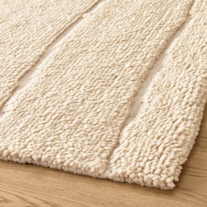 Könnte beinhalten: Ein cremefarbener Teppich mit einer dicken, hochflorigen und zotteligen Textur. Der Teppich besteht aus abwechselnd breiten, getufteten, strukturierten Bahnen, die durch schmale, glatte, flach gewebte Kanäle getrennt sind und ein Streifenmuster bilden.