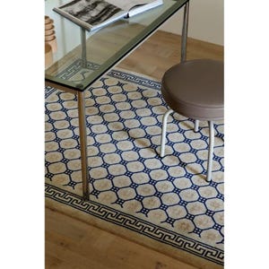 Könnte beinhalten: Ein rechteckiger Teppich mit cremefarbenem Hintergrund, einem sich wiederholenden blauen geometrischen Gittermuster und einer dunkelblauen griechischen Mäanderbordüre. Der Teppich liegt unter einem Schreibtisch mit Glasplatte und Metallbeinen.