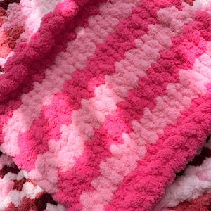 May include: A hand-knitted blanket featuring a thick, plush texture with alternating horizontal stripes in various shades of pink, ranging from light pastel pink to vibrant magenta and deep rose, creating a soft and cozy appearance throughout.