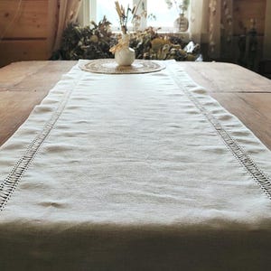 May include: A rectangular off-white table runner featuring a woven fabric texture and decorative openwork hemstitch detailing along both long edges. The runner is positioned lengthwise on a wooden surface, extending toward a small white vase.