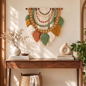 May include: A wall hanging made of macrame cord in shades of sage green, mustard yellow, terracotta, and cream. The piece features a wooden dowel, layered semicircular patterns, wooden beads, and several leaf-shaped fringe elements.
