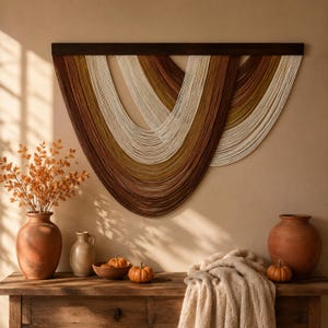 Könnte beinhalten: Ein Wandbehang mit mehreren Lagen drapiertem Garn in den Farben Creme, Senfgelb und Dunkelbraun, der an einer horizontalen Leiste aus dunklem Holz befestigt ist. Das Garn bildet überlappende U-förmige Kurven und erzeugt ein strukturiertes Bild.