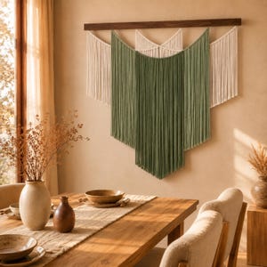 Peut inclure: Une tenture murale composée de cordes en macramé superposées en vert sauge et blanc cassé, suspendue à une tige en bois horizontale. Le design présente plusieurs sections en forme de V de franges pendantes, créant une œuvre d'art textile texturée.
