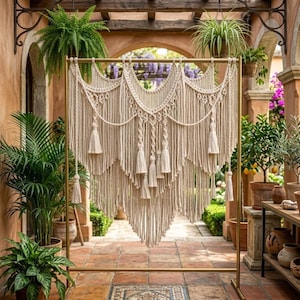 May include: A large off-white macramé wall hanging featuring intricate knotted patterns, draped cords, and multiple hanging tassels. The textile piece is suspended from a horizontal gold metal rod, showcasing a layered, symmetrical design with long fringe.