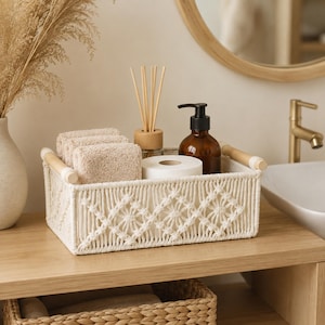 Può includere: Un cestino portaoggetti rettangolare in macramè color bianco sporco con motivo a rombi, due manici in legno chiaro e base solida. Il cestino contiene tre asciugamani beige piegati, un rotolo di carta igienica bianca e un dispenser di sapone.