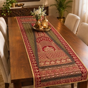 Könnte beinhalten: Ein langer, rechteckiger Tischläufer mit einem tiefroten und olivgrünen Webmuster, das komplexe florale und architektonische Bogenmotive zeigt. Der Stoff hat eine goldfarbene Bordüre und ist mit kleinen, wiederkehrenden Blumenmustern verziert.