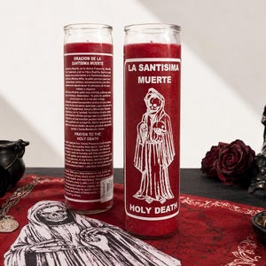 May include: A tall cylindrical red wax candle in a clear glass container featuring a white graphic of a hooded skeleton figure and the text La Santisima Muerte and Holy Death. The back of the glass displays a printed prayer in Spanish and English.