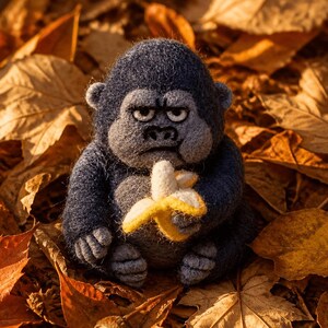 May include: A needle-felted dark gray gorilla figurine with a grumpy facial expression, holding a small yellow and white banana. The gorilla has a rounded body, textured wool-like surface, and small ears, sitting upright among fallen autumn leaves.