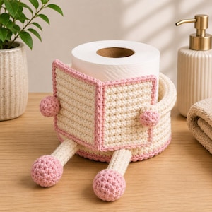 May include: A cream-colored crochet toilet paper holder featuring a pink-trimmed book design on the front, two extended legs with pink spherical feet, and a matching pink-trimmed circular base that holds a standard roll of white toilet paper.