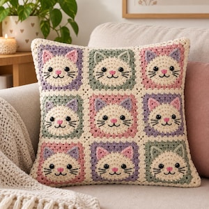 Pode incluir: Uma almofada quadrada de crochê com uma grade de nove rostos de gato. Cada gato é de cor creme com olhos de botão pretos, nariz bordado em rosa e bigodes pretos. Os gatos são emoldurados por quadrados em tons pastel.