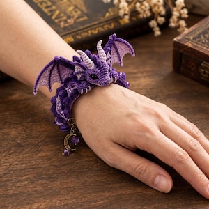 May include: A purple crocheted dragon bracelet featuring a detailed head with small horns, textured wings, and a coiled body. The bracelet includes a dangling bronze-toned charm shaped like a crescent moon with a small purple bead attached.