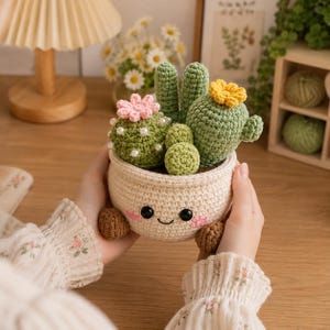 May include: A crocheted cactus arrangement in a cream-colored pot with a smiling face, pink cheeks, and two small brown feet. The pot contains three green crocheted cacti, one with white pearl accents and a pink flower, and one with a yellow flower.