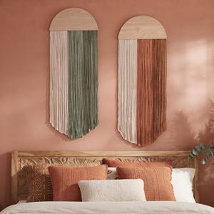 Può includere: Due decorazioni da parete con cime in legno chiaro semicircolari e lunghi cordoni verticali. Il pezzo a sinistra ha cordoni color crema e verde salvia, quello a destra color crema e terracotta, disposti in uno stile a frange stratificate.